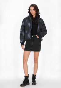 Zwarte pufferjack met een glanzende, gestructureerde afwerking, gecombineerd met een zwarte denim rok en zwarte veterschoenen.