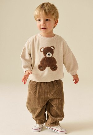 Young child wearing a beige sweatshirt with a brown teddy bear design, brown corduroy pants, and white sneakers, standing on a plain background.