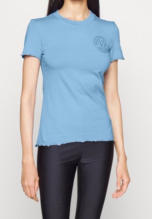 Kobieta w dopasowanym jasnoniebieskim t-shircie z krótkim rękawem z małym logo na piersi oraz czarnych legginsach.