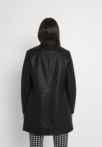 Abrigo de cuero negro con corte recto, cuello alto y sin herrajes visibles. Acabado texturizado. Llevado sobre pantalones de patrón en blanco y negro.