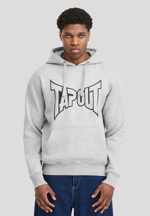 Mężczyzna w jasno-szarej bluzie z kapturem z napisem "TAPOUT" i niebieskich dżinsach, stojący na tle jednolitego szarego tła, patrzący przed siebie z neutralnym wyrazem twarzy.