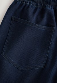 Materialul de culoare bleu marin are o textură moale, ribbed, cu un elastic la talie și un buzunar central pe spate, accentuat de cusături curate.