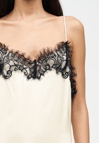 Γυναίκα που φοράει εκρού σατέν καμισόλ με λεπτή μαύρη δαντέλα κατά μήκος της γραμμής του ντεκολτέ και λεπτές ράντες στους ώμους.