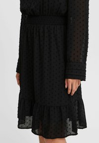 Robe noire en tissu texturé avec de petits points en relief, taille smockée, manches longues et ourlet festonné. Les manches semi-transparents ajoutent du détail.