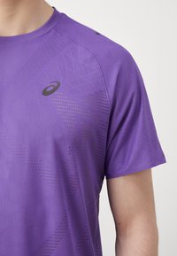 Lila Sportshirt mit kurzen Ärmeln, bietet ein strukturiertes geometrisches Muster, atmungsaktives Material und ein kleines dunkles Logo auf der Brust.