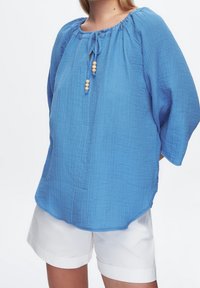 Blusa leggera blu con tessuto texture, scollatura arricciata e chiusura a fiocco decorata con perline di legno. Maniche corte e leggermente a sbuffo.