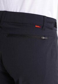 Gros plan d'un pantalon noir avec une poche arrière zippée et une petite étiquette en tissu rouge près de la taille, porté avec une chemise blanche.