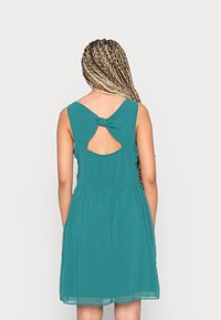 Vestido azul-petróleo com corpete justo e saia evasê. Apresenta um recorte nas costas único e um detalhe de laço. Feito de um tecido leve e fluido.
