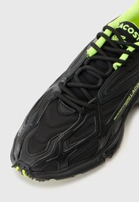 Fekete sneaker, textúrált felsőrésszel, neon zöld részletekkel és karcsú dizájnnal. Fekete fűzők és márkajelzés az oldalon.