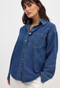 Camisa de mezclilla en azul oscuro con cierre de botones en la parte delantera, cuello y un bolsillo en el pecho. Presenta un corte relajado y detalles de costura visibles.