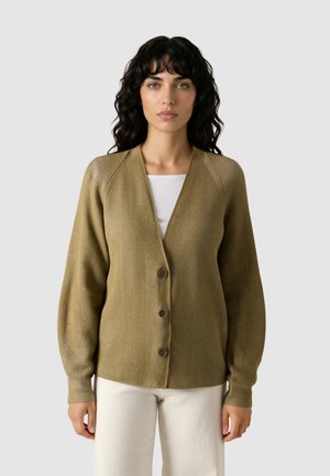 Vrouw met golvend donker haar, gekleed in een olijfgroene, dichtgeknoopte cardigan over een wit shirt en lichtgekleurde broek, staand tegen een effen achtergrond.