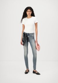Camiseta ajustada de manga corta blanca, jeans skinny azul claro con un pañuelo negro atado a la cintura, bolso de mano rosa y zapatos planos negros con detalles.