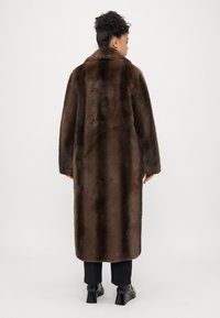 Hnedý umelý kožušinový dlhý kabát s golierom, ktorý má mäkkú textúru a vertikálny pruhovaný vzor. Nosený cez elegantné čierne nohavice a topánky.