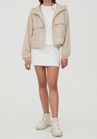 Chaqueta beige con capucha, dos bolsillos frontales, puños elásticos, llevada sobre una camisa blanca de manga corta y una falda de mezclilla blanca, combinada con botas blancas.