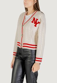 Cardigan in maglia a trecce crema con accenti a strisce rosse, grande ricamo "NY" e bottoni in legno. Indossato sopra pantaloni in pelle neri.