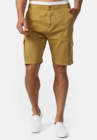 Mand iført sennepsfarvede cargo shorts med forlommer og hvide slip-on sko, stående mod en ensfarvet baggrund.