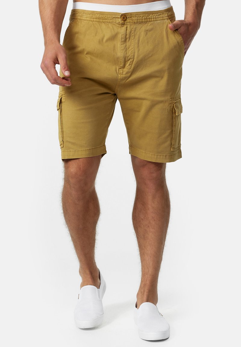 Mand iført sennepsfarvede cargo shorts med forlommer og hvide slip-on sko, stående mod en ensfarvet baggrund.