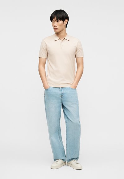 Jeune homme debout, les mains dans les poches, portant un polo beige à manches courtes, un jean large bleu clair et des baskets blanches sur fond uni.