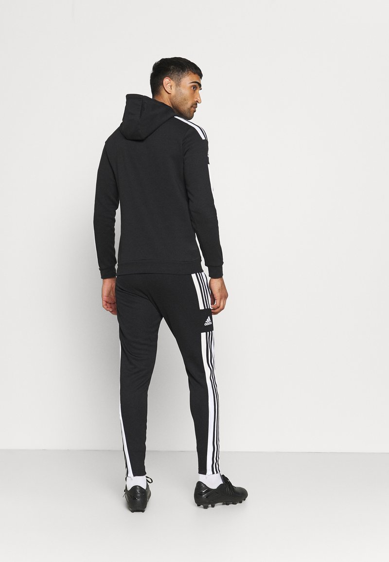 Haut à capuche noir avec une capuche et des poignets côtelés, associé à un jogger noir avec des rayures blanches et un logo Adidas sur la jambe.