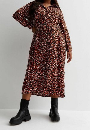 Mujer que lleva un vestido midi de manga larga con estampado marrón y negro, con un lazo en la cintura y botas negras gruesas hasta el tobillo, de pie frente a un fondo liso.