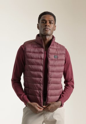 Chaleco de plumas burdeos con diseño acolchado, cuello alto y cremallera frontal, usado sobre un suéter a juego, combinado con pantalones beiges.