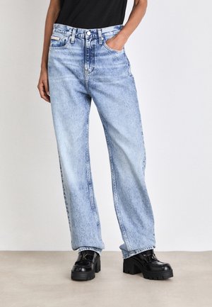 Osoba ubrana w jasnoniebieskie, dopasowane w talii jeansy o prostych nogawkach oraz czarne masywne buty na platformie, stojąca z jedną ręką w kieszeni.