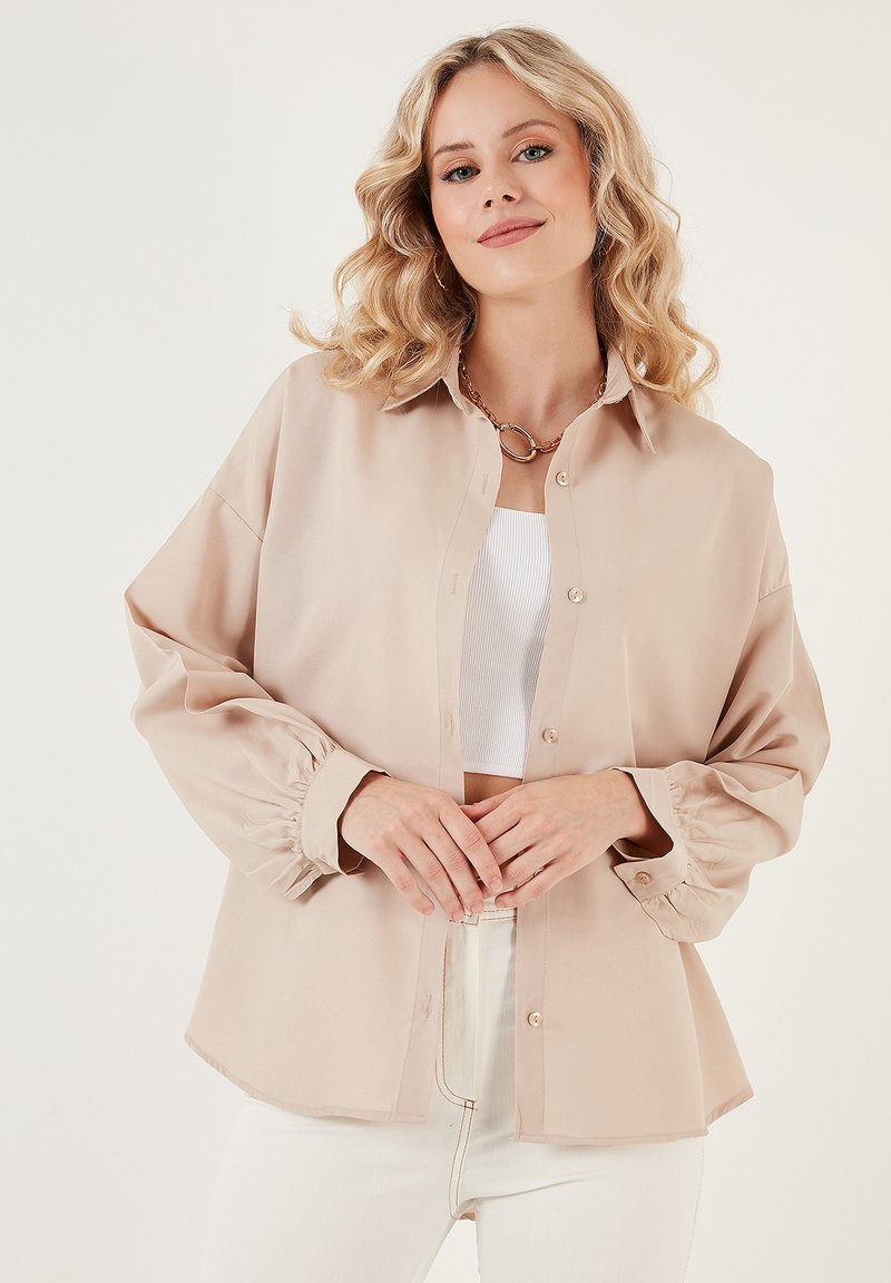 Camicia beige con bottoni realizzata in tessuto setoso, caratterizzata da un taglio morbido, polsini elasticizzati e orlo arrotondato, indossata sopra una maglietta bianca.