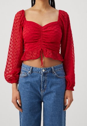 Vrouw draagt een rood gerimpeld cropped topje met bloemmotief doorzichtige pofmouwen en een high-waisted blauwe spijkerbroek, staand tegen een witte achtergrond.