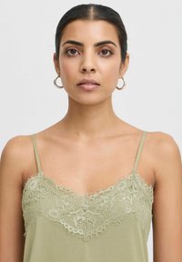 Camisola verde con pizzo decorativo sul collo, spalline sottili e morbidezza al tatto. Presenta dettagli di pizzo sfrangiato lungo i bordi.