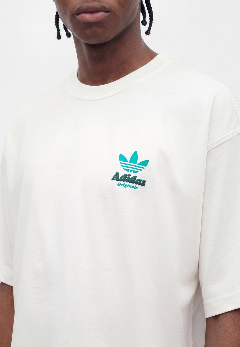 Junge Person mit geflochtenem Haar, die ein weißes Adidas Originals T-Shirt mit blauem Logo auf der Brust vor einfarbigem Hintergrund trägt.