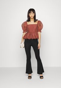 Blusa de mangas bufantes em cor ferrugem com decote quadrado e bainha peplum, combinada com calças pretas de cintura alta. A modelo segura uma pequena bolsa branca.