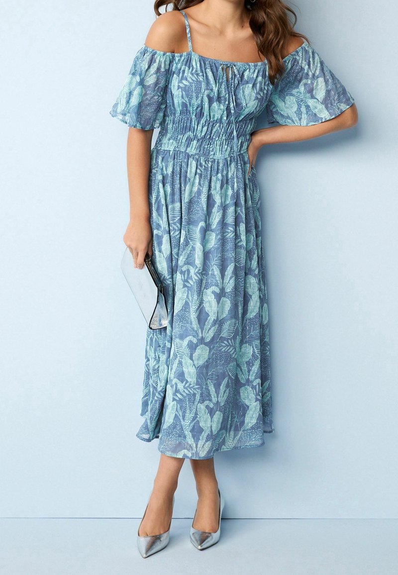 Vestido maxi de estampado floral azul con mangas caídas, cintura fruncida y falda fluida. Clutch metálico y tacones puntiagudos plateados.