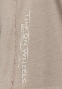 Camiseta de algodón beige con el texto blanco "VIDA EN RUEDAS" y "PISTA RÁPIDA" en vertical a lo largo del lado, con un detalle de costura roja contrastante.