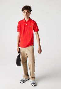 Polo rojo de manga corta y cuello, complementado con pantalones beiges con franjas laterales verdes y un bolso negro con asa.