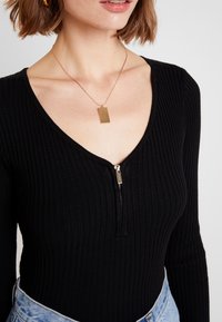 Zwart geribbelde bodysuit met lange mouwen, voorzien van een V-hals en een ritssluiting aan de voorkant. Gecombineerd met een gouden rechthoekige hangersketting.