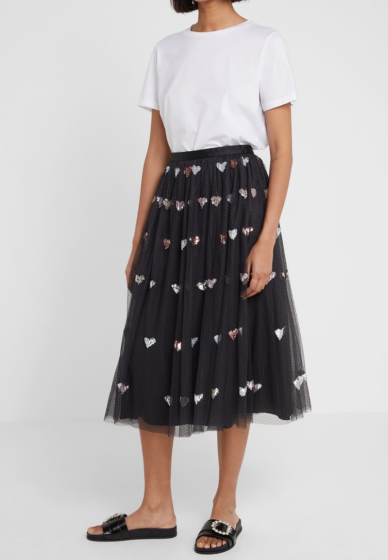 Femme portant une chemise blanche à manches courtes, une jupe midi en tulle noire ornée de cœurs en sequins et des sandales noires, à brides, avec une boucle en perles, vue de profil.