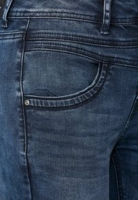 Mörkblå jeans i denim med synlig sömnad, en ficka med en metalldorn och en något urblekt textur. Närbild av baksidan.