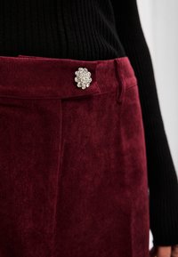 Burgundijas korduroja svārki ar teksturētu virsmu, kuriem ir sudraba dekoratīvā pogas ar ziedu dizainu jostasvietā.