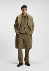 Beige uldfrakke over en olivengrøn kordfløjlsk jacket og matchende bukser. Sorte sko med en chunky sål. Minimalistisk design, glatte teksturer.