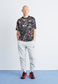 Sort T-shirt med trykte billeder, grå sweatpants og sorte sneakers med røde accenter. Outfittet har forskellige teksturer og mønstre.