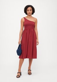 Vestido de línea A de un solo hombro en un profundo tejido rojo, con un corpiño ajustado y un cinturón fruncido. Combinado con un bolso texturizado en azul marino y sandalias negras.