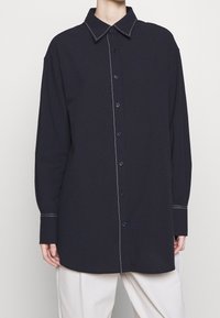 Chemise bleu marine à boutons avec un col classique, des manches longues et des surpiqûres blanches contrastantes le long du devant et des poignets.