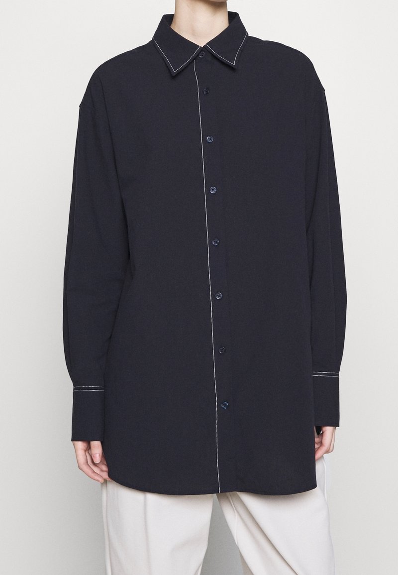 Chemise bleu marine à boutons avec un col classique, des manches longues et des surpiqûres blanches contrastantes le long du devant et des poignets.