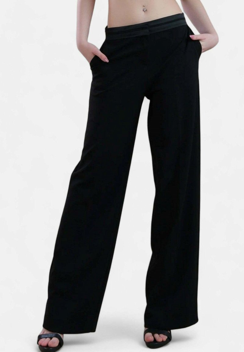 Personne portant un pantalon noir taille haute à jambes larges, mains dans les poches, et des sandales noires à bouts ouverts sur un fond blanc.