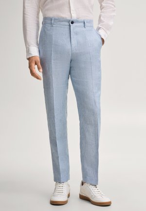 Man draagt lichtblauwe op maat gemaakte broek, wit overhemd met lange mouwen en witte sneakers, staand tegen een effen achtergrond.
