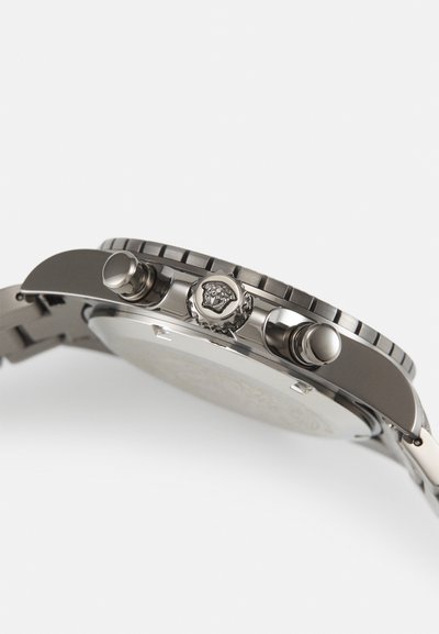 Stainless steel watch with a round case, textured bezel, and crown featuring a lion emblem. Two pushers adjacent to the crown.