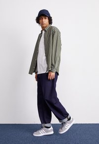 Giovane uomo con capelli ricci che indossa un berretto blu navy, una camicia a righe verdi sopra una maglietta bianca, jeans scuri con risvolti e sneaker grigie Adidas, in piedi su un tappeto blu.