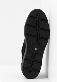 Semelle de chaussure noire avec des rainures profondes et un petit logo circulaire près du centre, positionnée verticalement sur un fond blanc.