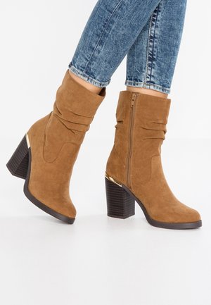 Brune ruskindsstøvler til ankler med blokhæle og lynlåse på siden, båret sammen med blå skinny jeans.