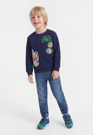 Garçon blond souriant debout, portant un sweat-shirt bleu marine avec des motifs de dinosaure et de chien, un jean bleu et des baskets vertes et bleues.